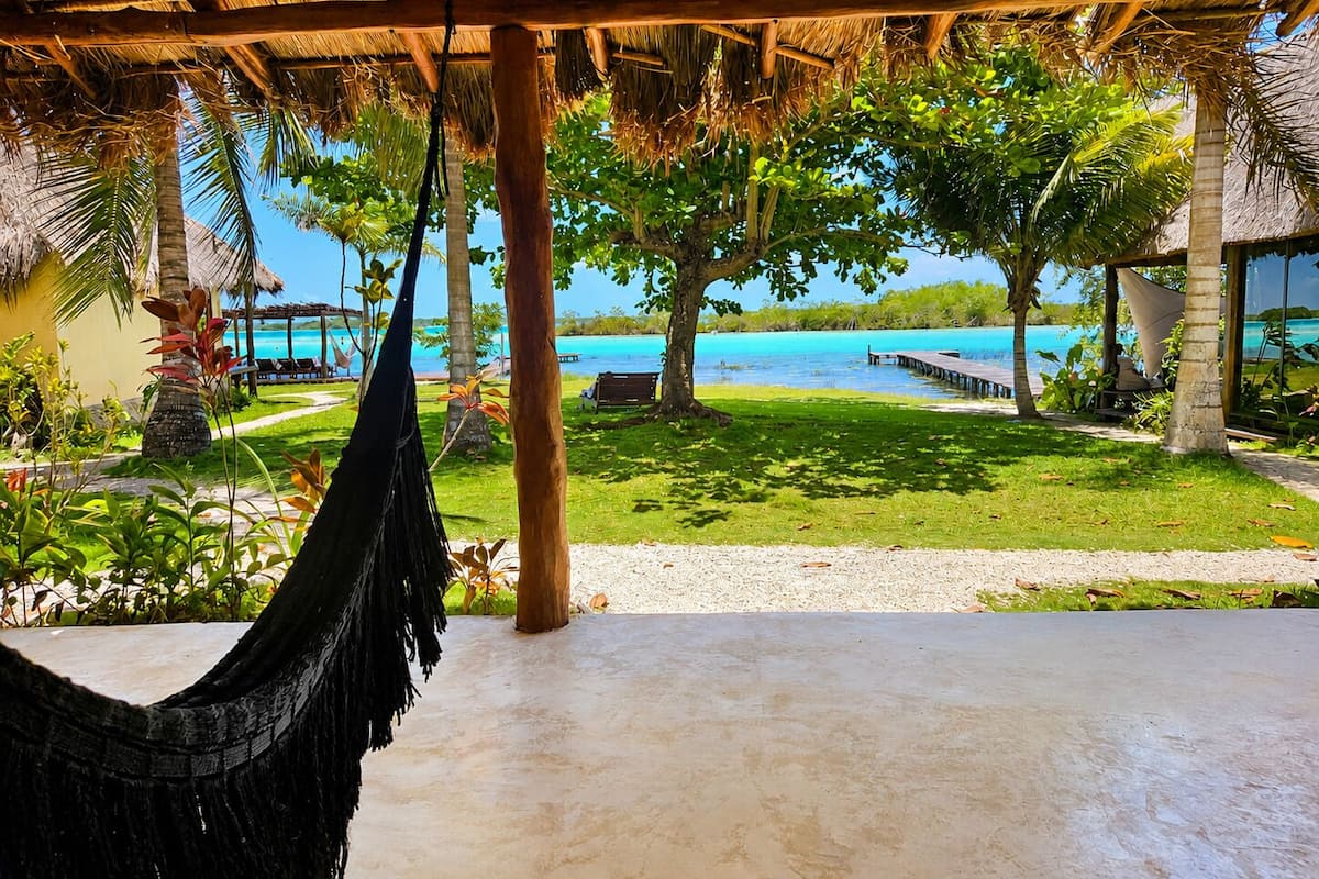 A hammock under a thatched roof, a view of a beach, and a clear blue sky.
