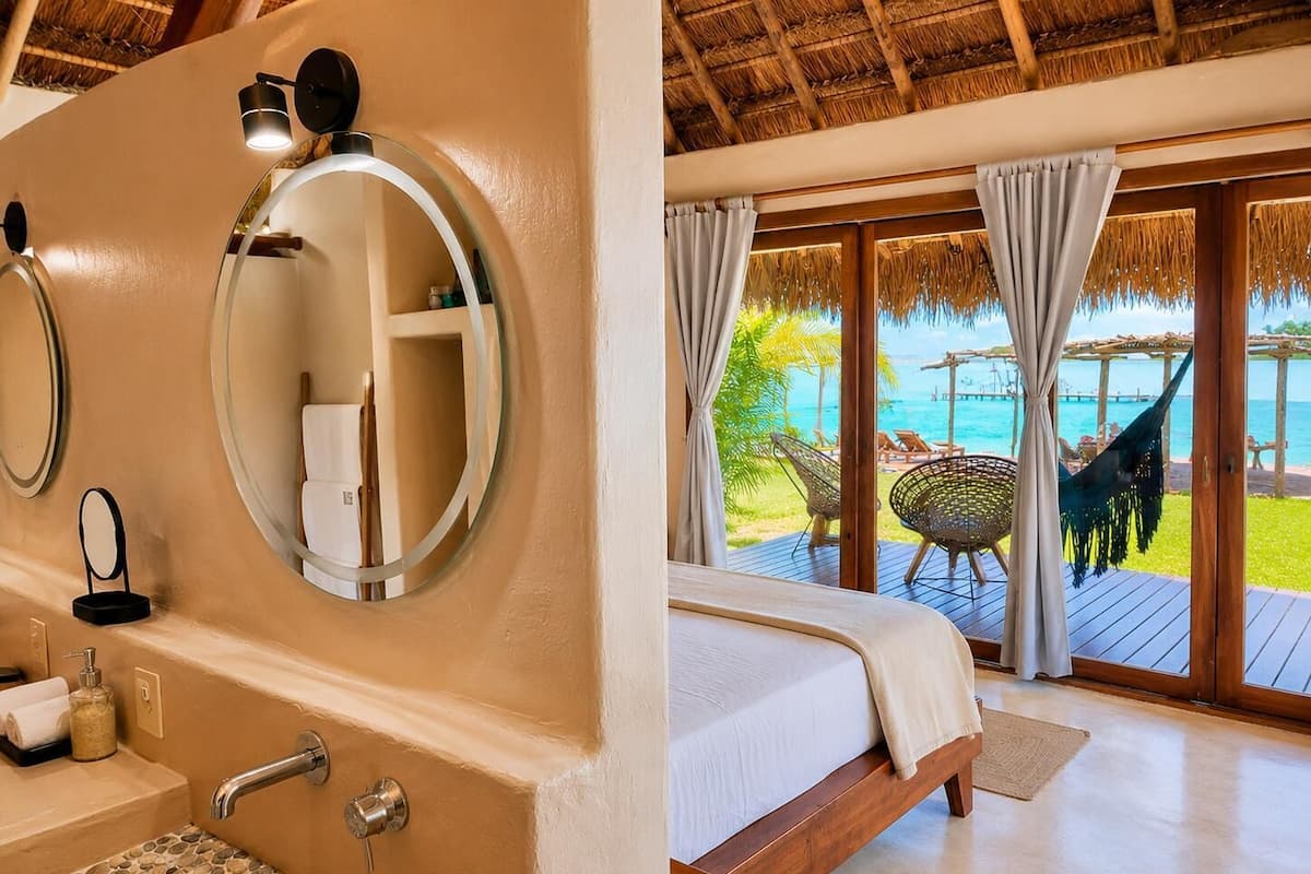 A bathroom with a round mirror, a sink with a mosaic tile design, and a toilet. The room has a wooden ceiling and a large window with curtains.