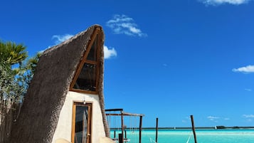 A thatched-roof hut with a wooden deck, two wicker chairs, and a wooden staircase leading to a door.