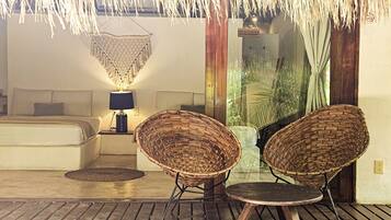 Une pièce comprenant un lit, une table de chevet avec une lampe, deux fauteuils en osier, une petite table et un plafond recouvert de chaume.
