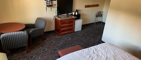 A hotel room with a bed, a desk with a TV, a chair, a small refrigerator, and a carpet with a circular pattern.