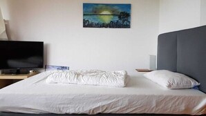 Una cama bien hecha con edredón blanco y almohadas, un televisor de pantalla plana sobre un atril de madera y un cuadro de un paisaje en la pared.