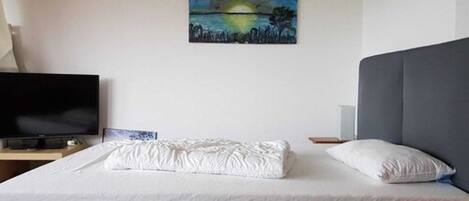A neatly made bed with a white comforter and pillows, a flat-screen TV on a wooden stand, and a painting of a landscape on the wall.