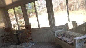 A sunroom with a table and chairs, a wicker sofa, and large windows overlooking a wooded area.