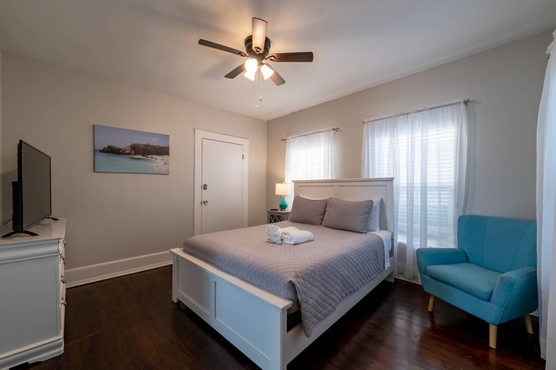 Un dormitorio con cama, ventilador de techo, televisión, un cuadro, una puerta, ventana con cortinas y una silla azul.