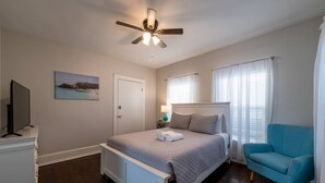 Un dormitorio con cama, ventilador de techo, televisión, un cuadro, una puerta, ventana con cortinas y una silla azul.