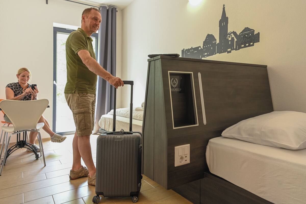 A hotel room with a bed, a desk, and a chair. There is a suitcase on the floor.