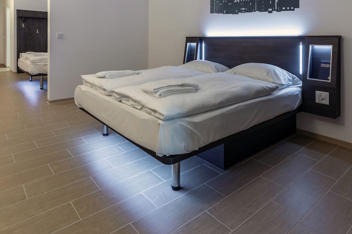 A modern hotel room with a large bed, a bedside table, and a wall-mounted control panel.
