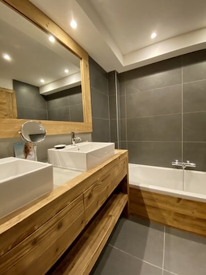 Een moderne badkamer met twee witte wasbakken, een houten wastafelmeubel, een grote spiegel en een bad.