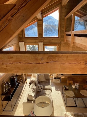Een gezellig chaletinterieur met een slaapzolder, houten balken, een zithoek, een eettafel en grote ramen met uitzicht op de bergen.