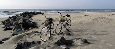 Deux vélos sont garés sur une plage de sable, avec une côte rocheuse et l’océan en arrière-plan.