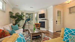 A living room with a white sofa, colorful cushions, a wooden coffee table, a peacock sculpture, a fireplace, and a television.