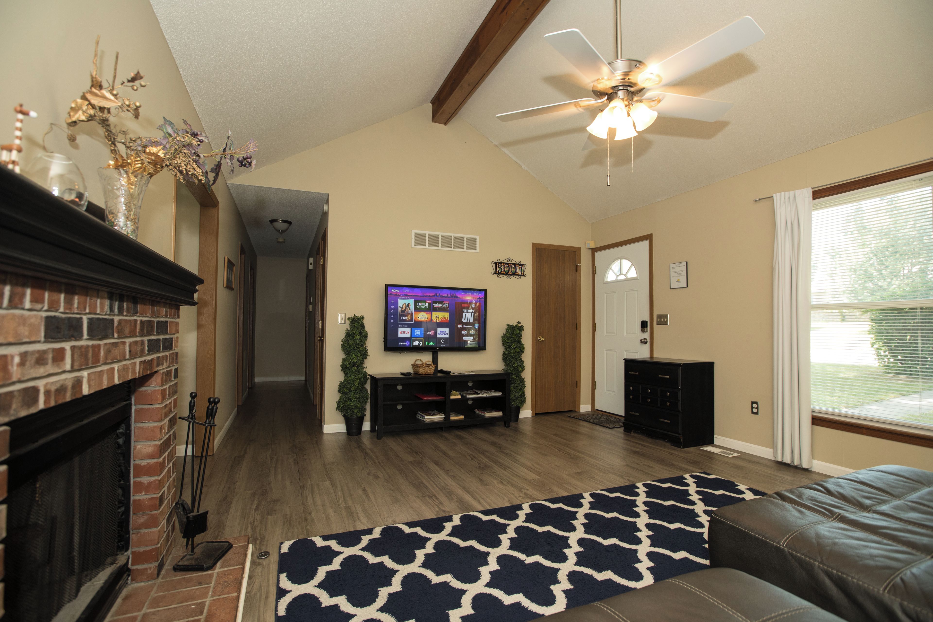 A living room with a fireplace, a television mounted on the wall, a ceiling fan, and a large window.