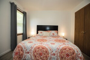 A bedroom with a bed, two bedside lamps, a window with curtains, and a wooden wardrobe.