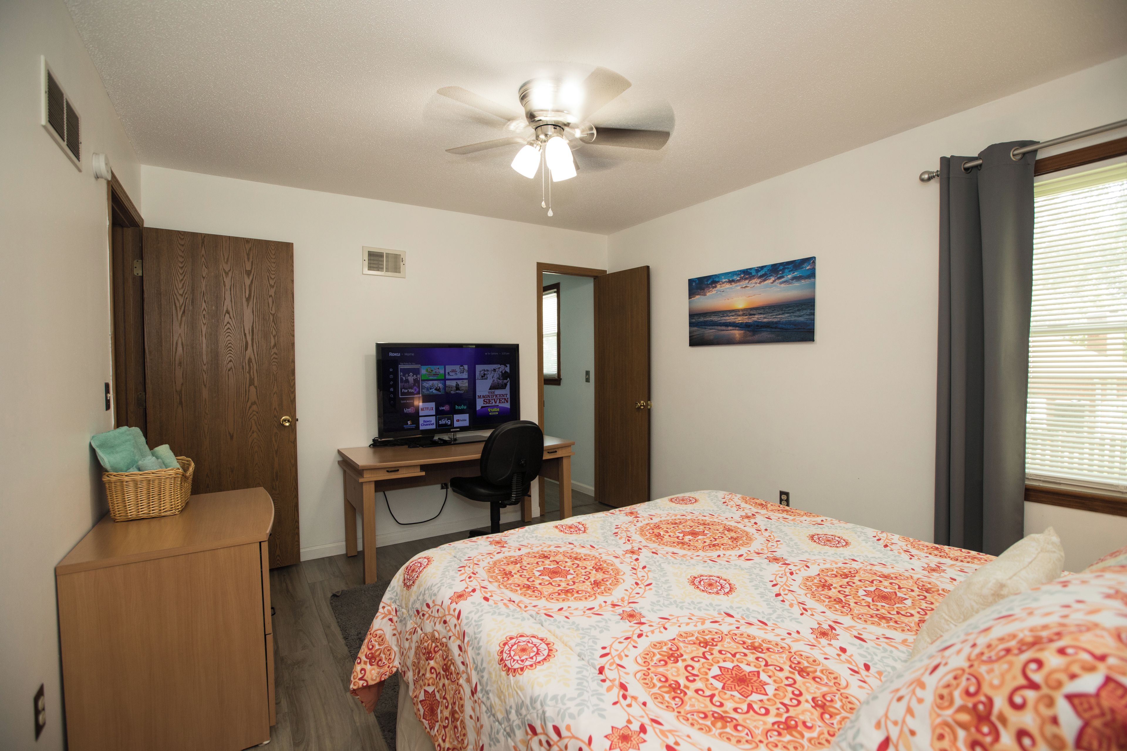 Quarto com cama, escrivaninha com computador, ventoinha no teto, janela com cortinas e quadro pendurado na parede.