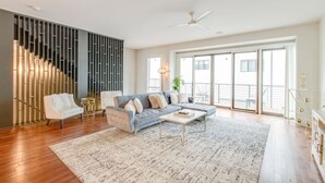 Ein geräumiges Wohnzimmer mit grauem Sofa, einem weißen Sessel und einem Couchtisch. Links befindet sich eine moderne Treppe, rechts ein Balkon mit Geländer.