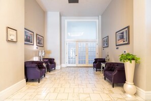A spacious hallway with a marble floor, flanked by purple armchairs and a tall white vase with green foliage.