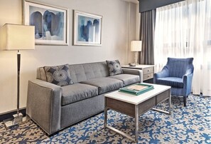 Ein Hotelzimmer mit einem grauen Sofa, einem passenden Sessel, einem kleinen Tisch, einem gemusterten Teppich und zwei gerahmten Kunstwerken an der Wand.
