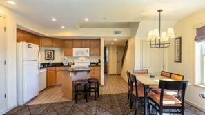 A modern kitchen with wooden cabinets, a white refrigerator, a microwave, and a stove. There is a dining area with a table and chairs. - Angels Camp | Friends & Family Golf Getaway | Pool + Golf | 3BR Sleeps 8 (Angels Camp)
