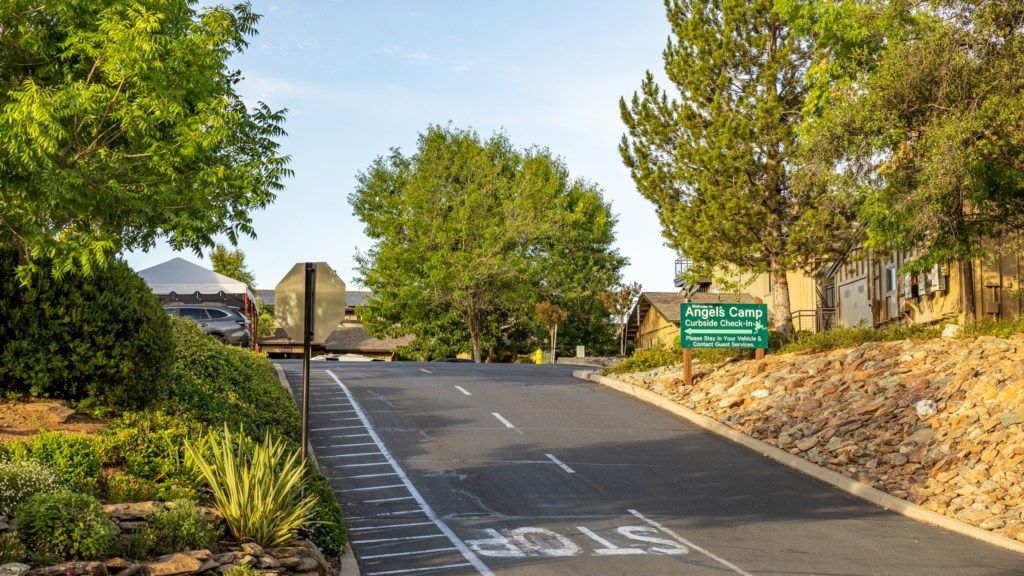 A road leading to a camp with a sign indicating 'Angels Camp' and a curbside check-in area.