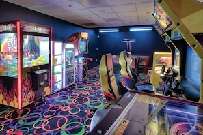 An arcade with various gaming machines, including claw machines and racing games, with a colorful floor design.