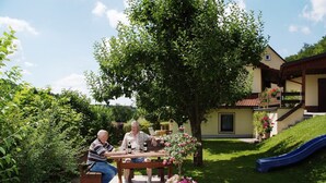 Dos personas sentadas en una mesa al aire libre, con un árbol y una casa al fondo.