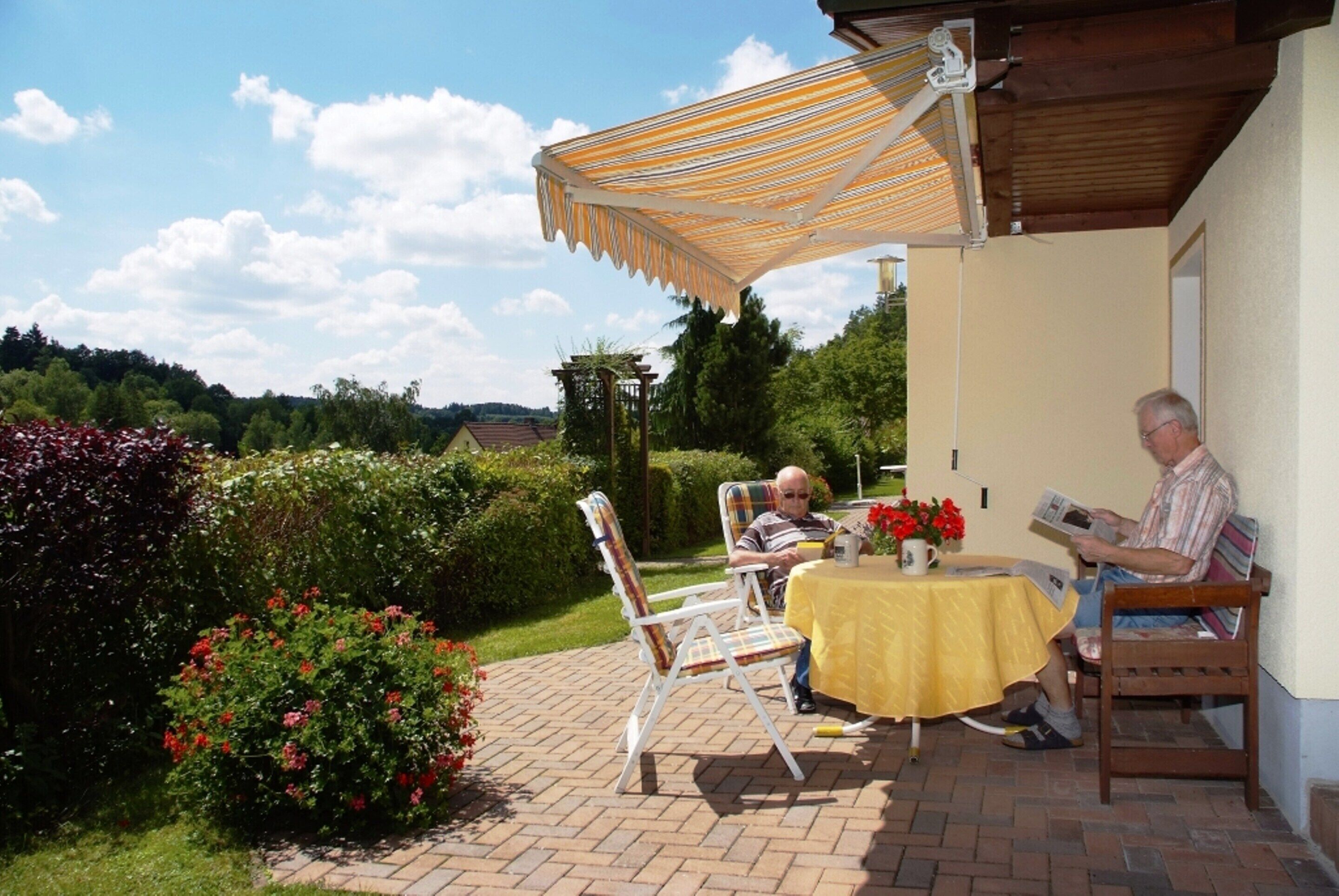 Een terras buiten met een gele tafel, twee stoelen en een gestreepte parasol.