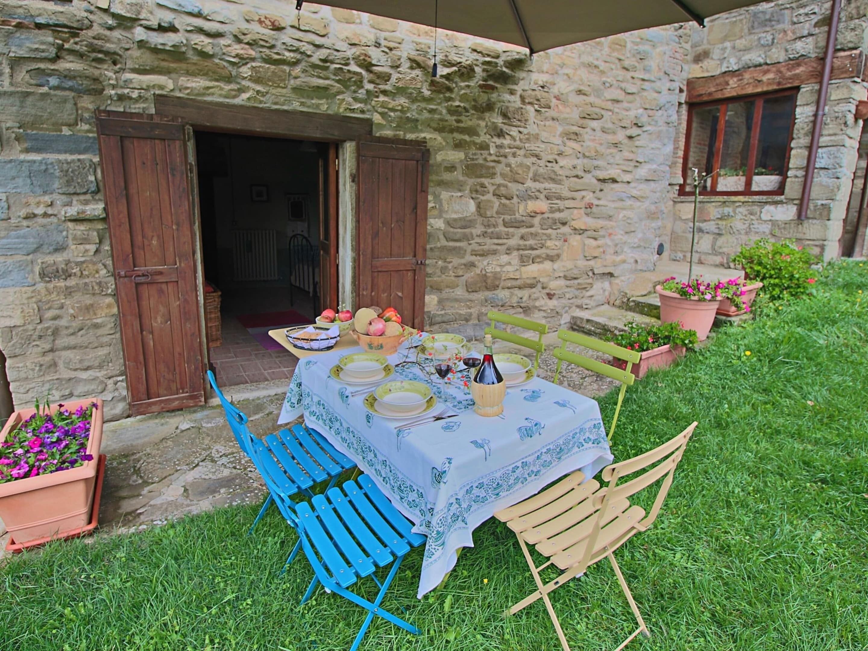 An outdoor dining area with a stone building, wooden chairs, and a table set with plates, bowls, and a bottle.