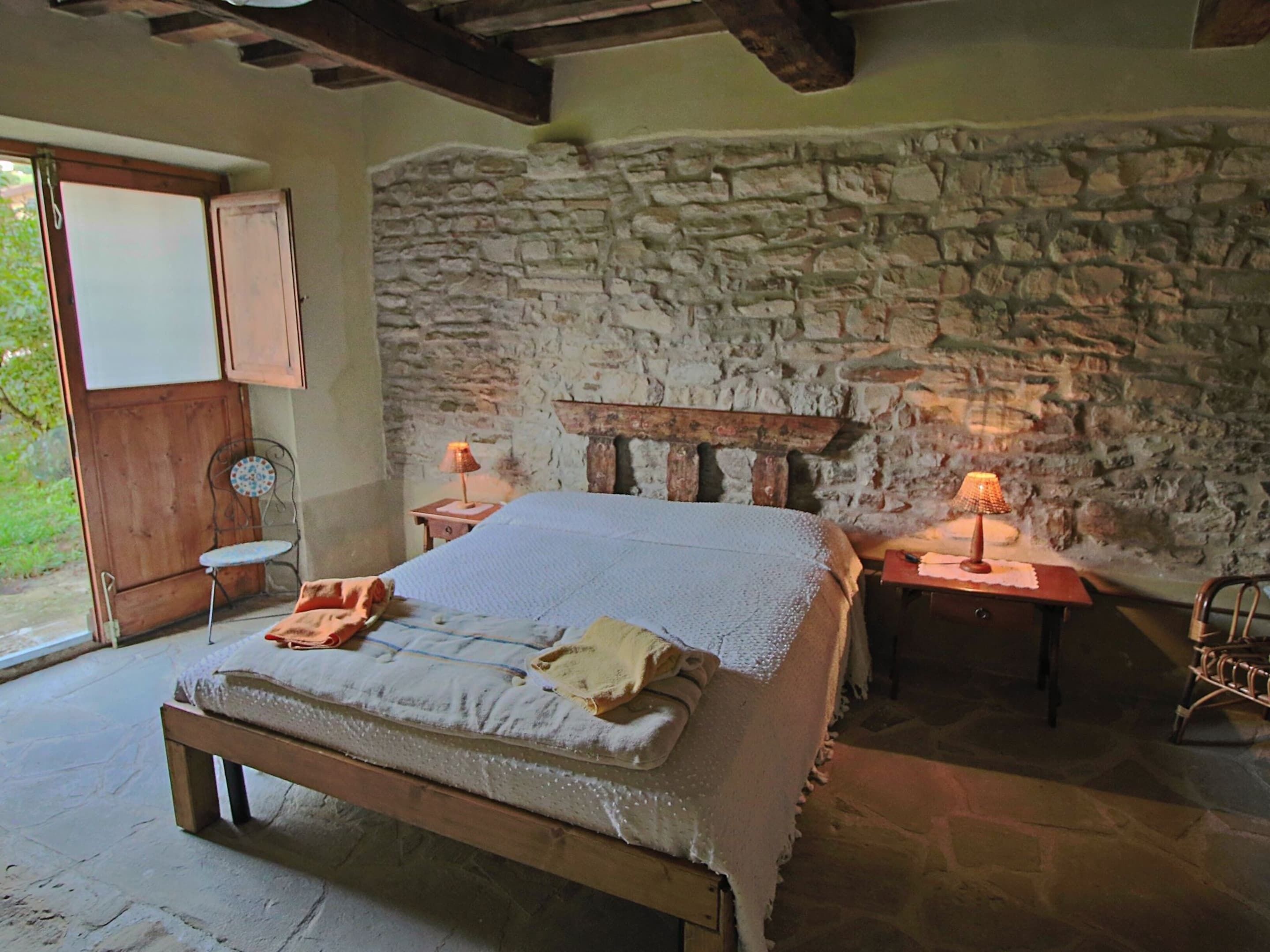 Un dormitorio rústico con pared de piedra, cama de madera, dos mesitas de noche con lámparas y una pequeña silla.