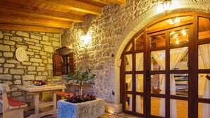 Un intérieur aux murs de pierre, avec une table en bois dressée pour le repas, un bac à fleurs en pierre orné de plantes et une grande porte en bois en arc de cercle donnant sur l’extérieur.