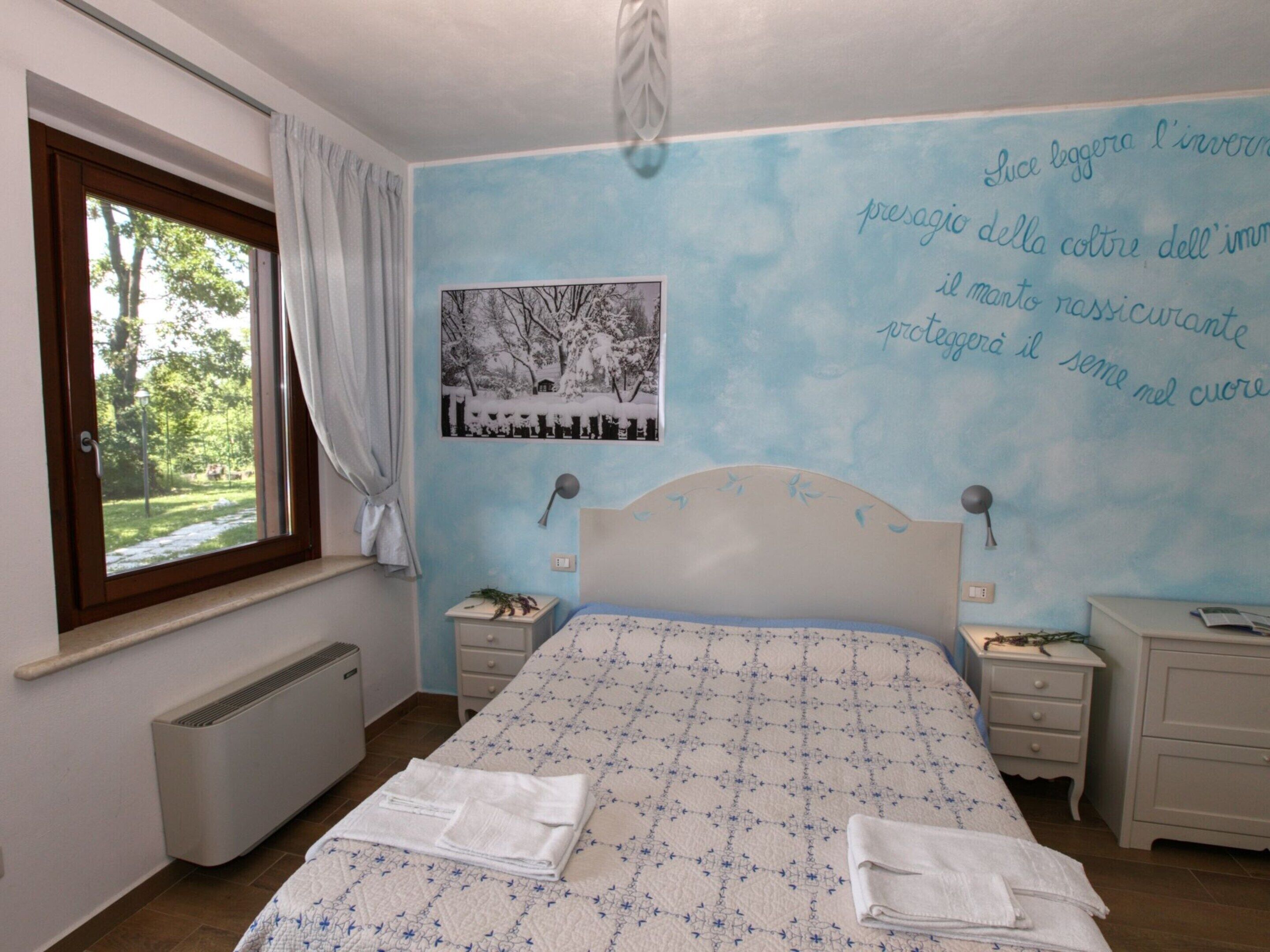 A bedroom with a bed, two bedside tables, a window with curtains, a radiator, and a wall with a framed picture and text.