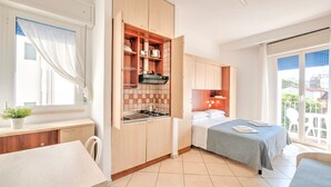 A compact, modern kitchen with wooden cabinets, a countertop, and a stove. Adjacent is a bedroom with a bed, a nightstand, and a window with curtains. - Modern Apartment in Rimini With Balcony (Rimini)