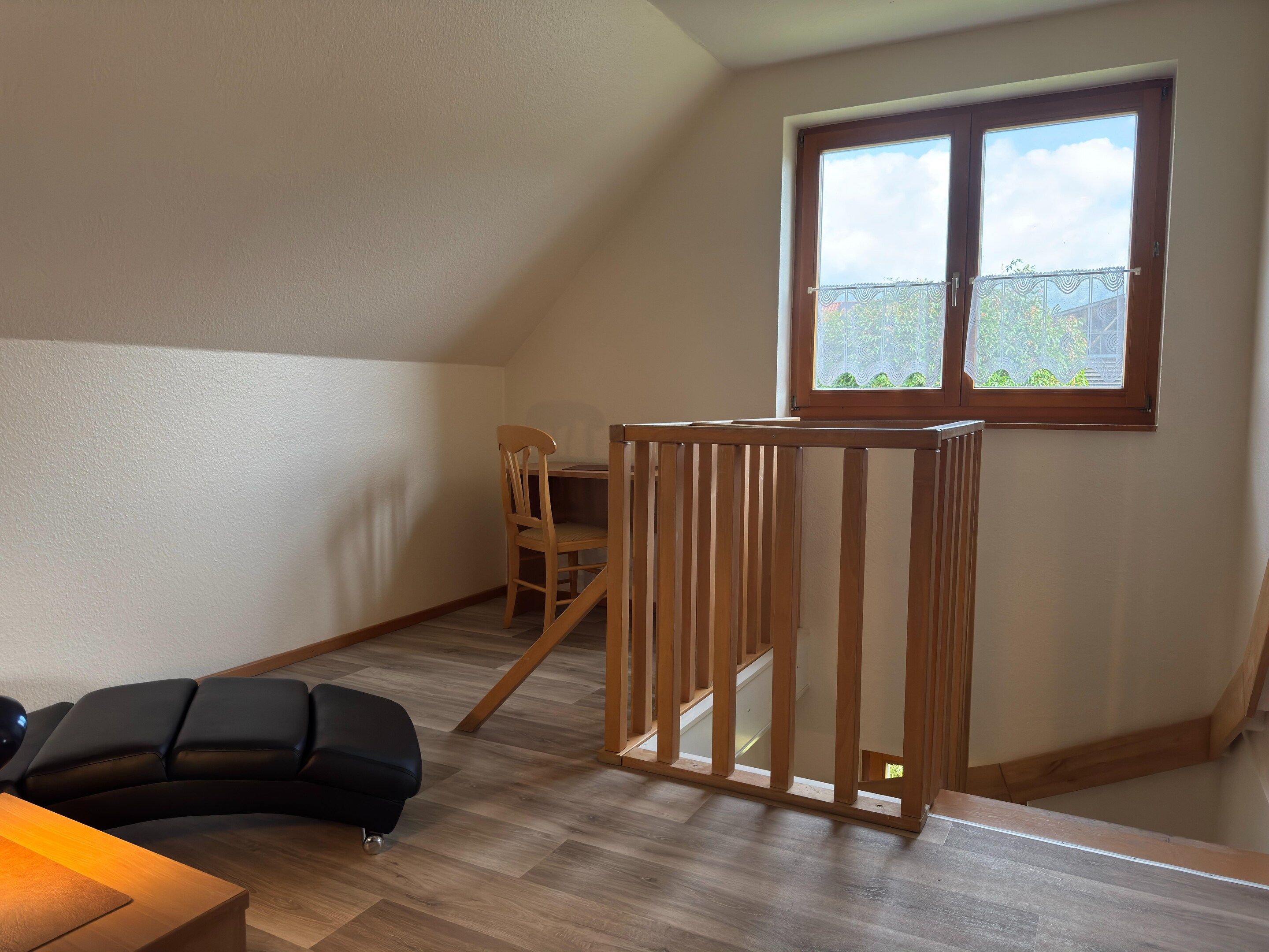 A room with a wooden desk, a black chair, a wooden railing, and a window with a view of greenery.