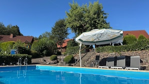 A swimming pool with a lounge area, a covered seating area, and a building with a red roof in the background. - Holiday Home in the Teutoburg Forest (Brakel)