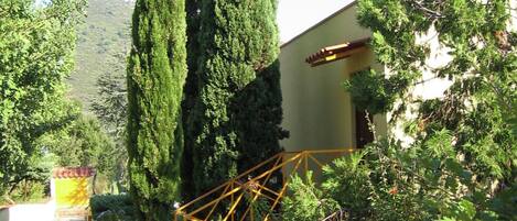 A building with a yellow facade, a staircase with a yellow railing, and surrounding greenery including tall trees and shrubs.