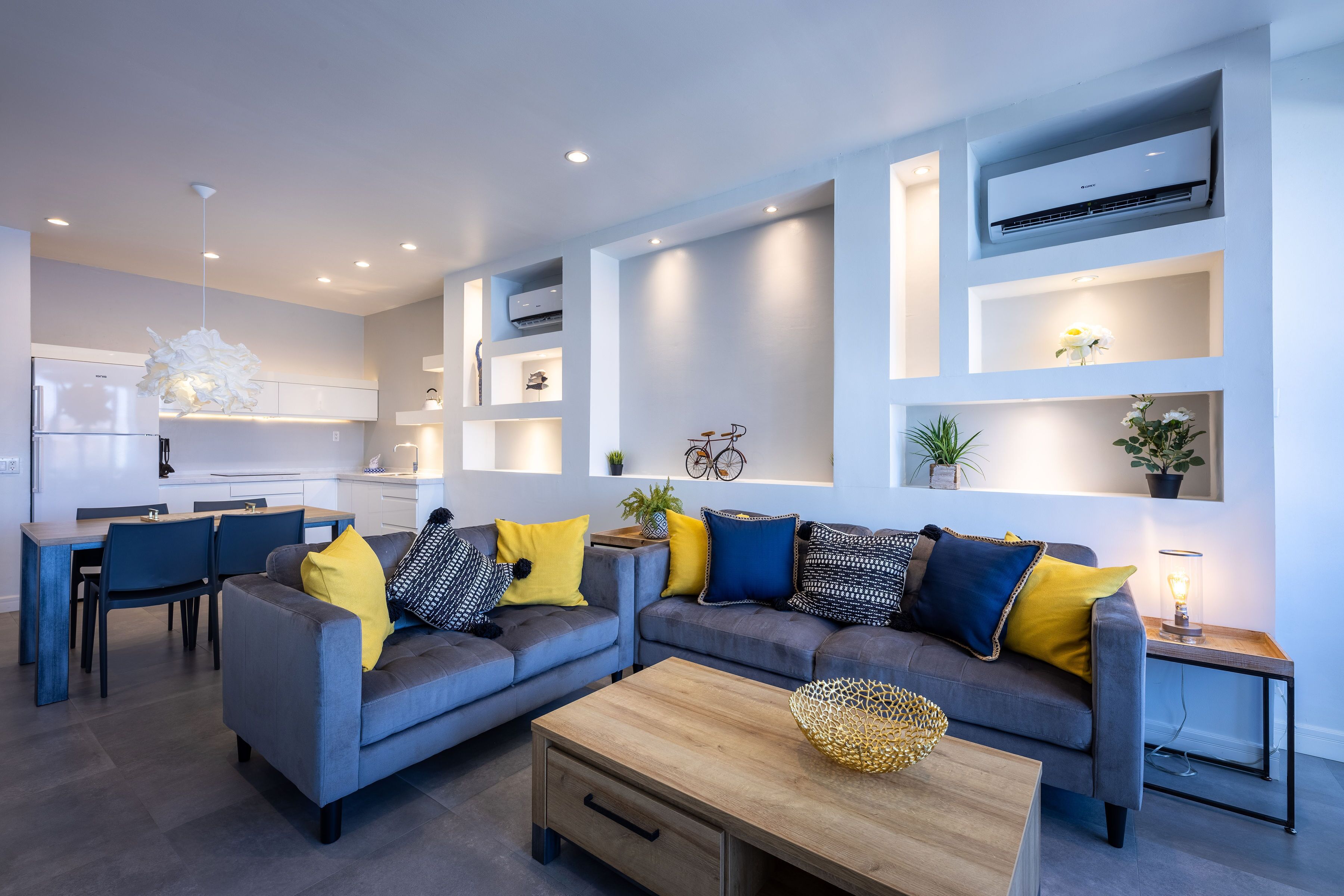 A modern living room with grey sofas, yellow and blue cushions, a wooden coffee table, and built-in shelving with decorative items.