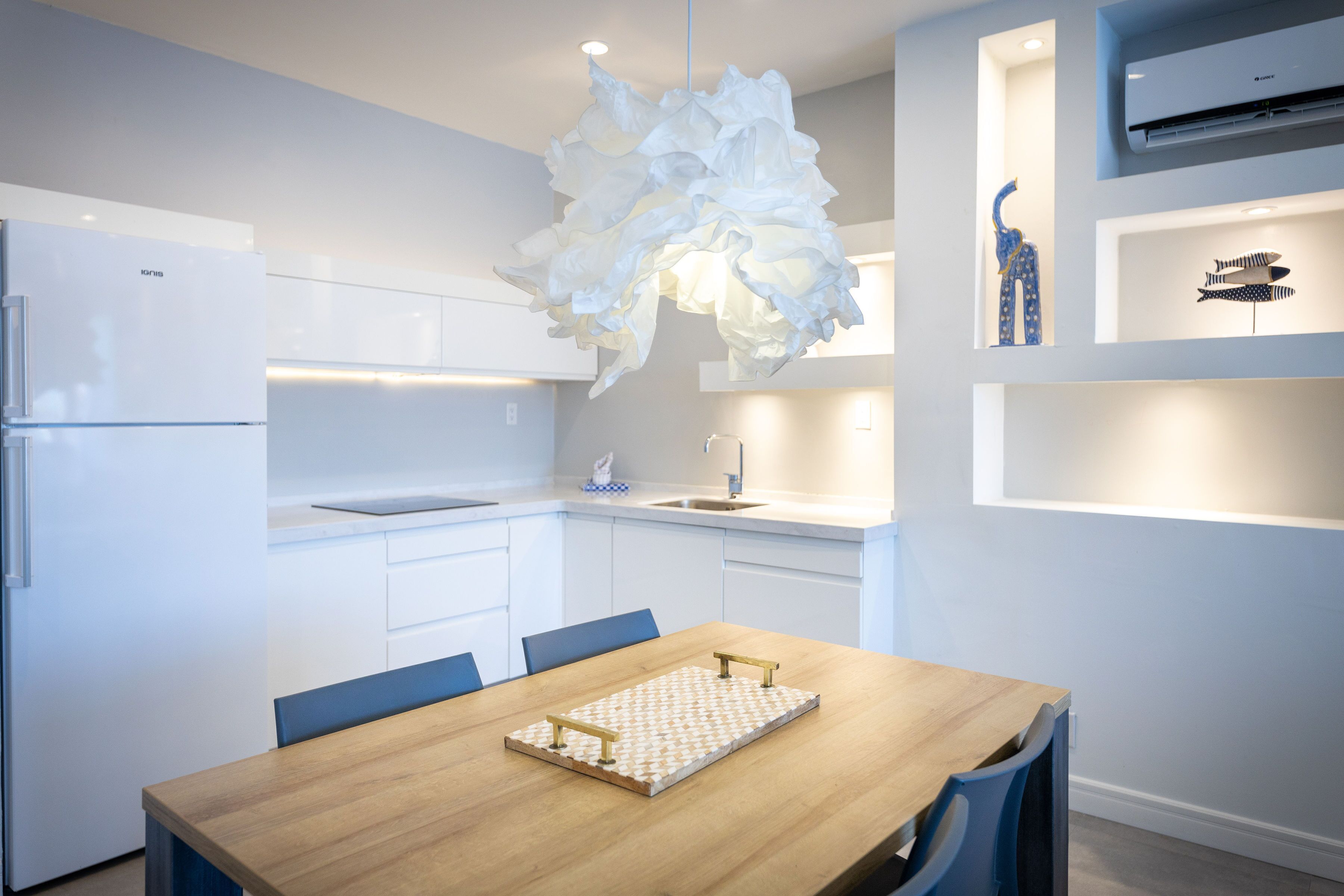 Une cuisine moderne avec des meubles blancs, une table à manger en bois et un lustre blanc original.