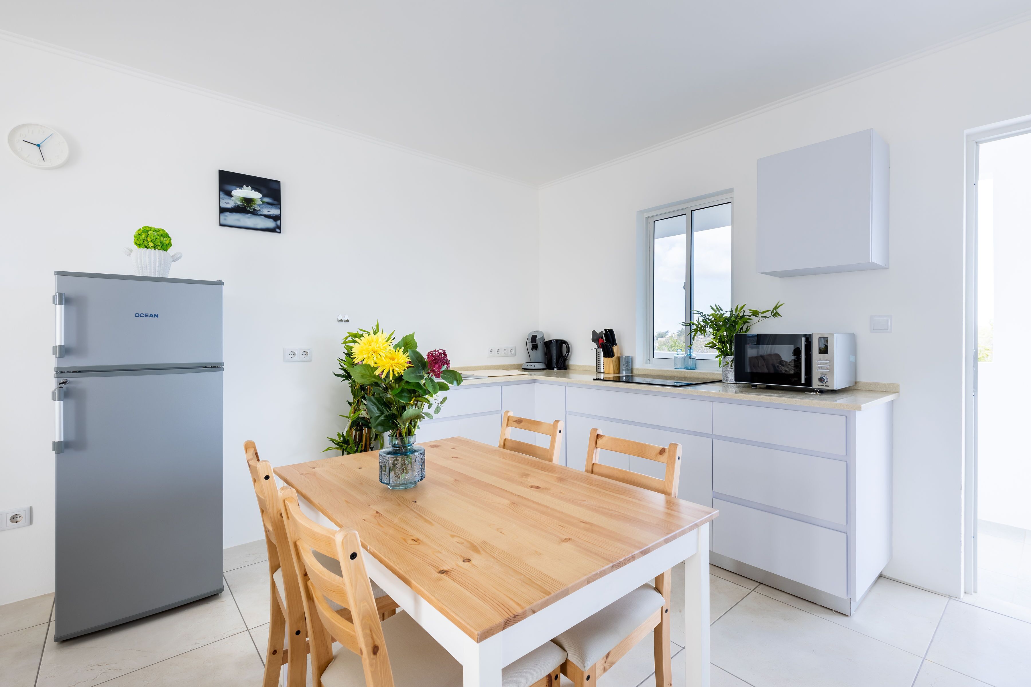 A modern kitchen with white cabinets, a grey refrigerator, a wooden dining table with chairs, and a vase of flowers.