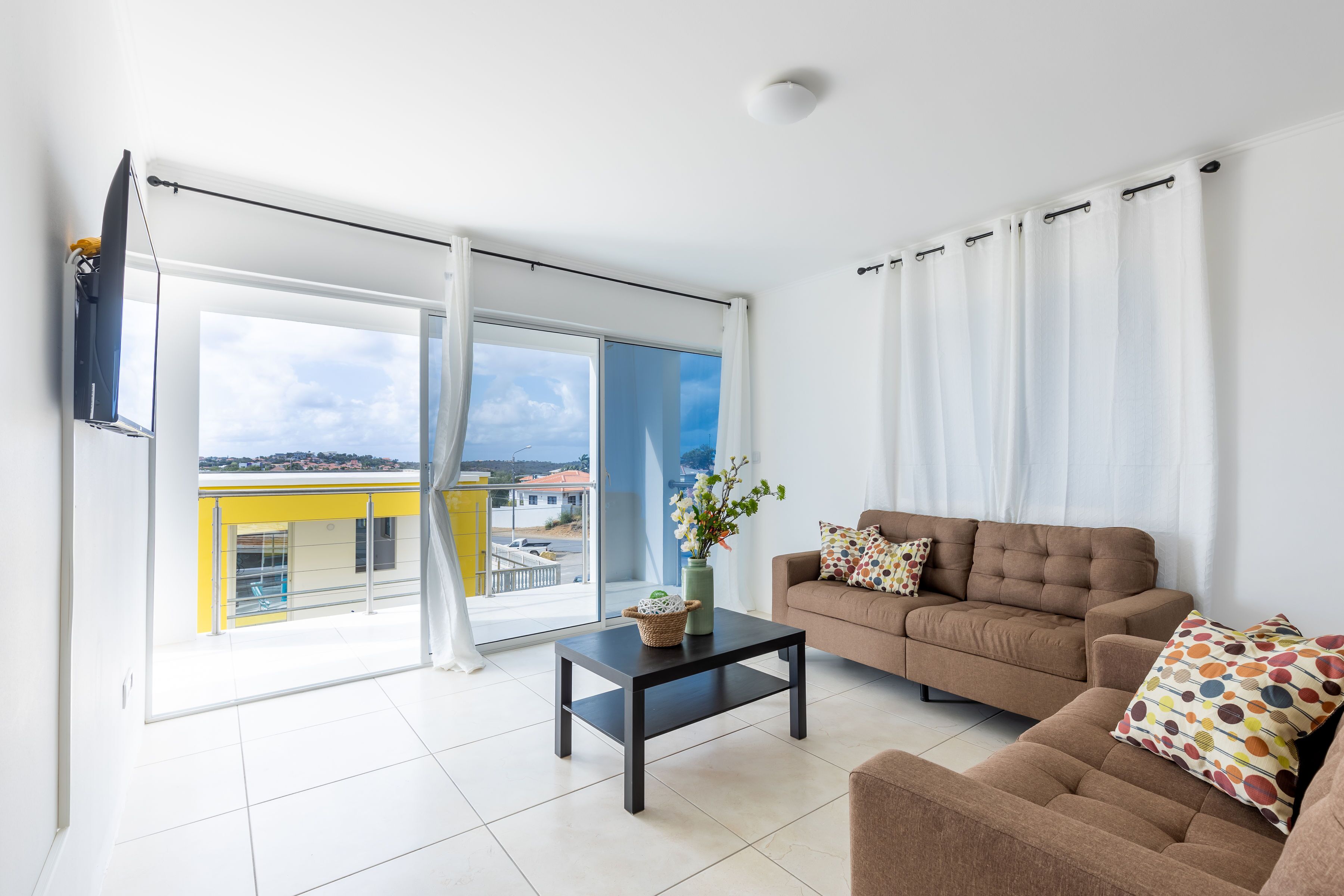 A modern living room with a sofa, a coffee table, and a view of a yellow building through large windows.