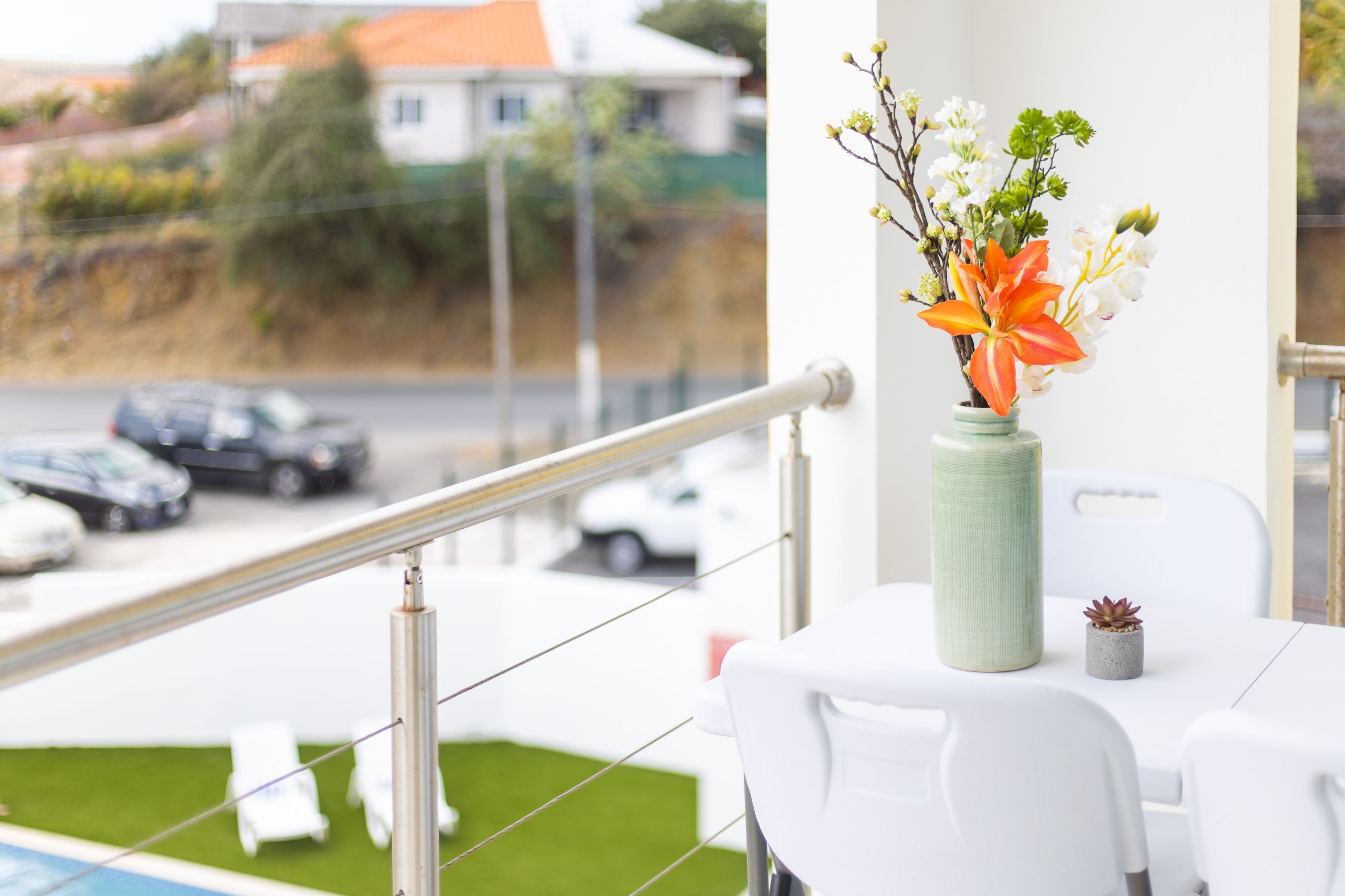 Ein Balkon mit Geländer, ein Tisch mit Blumenvasen und Blick auf eine Straße mit Autos und Häusern.
