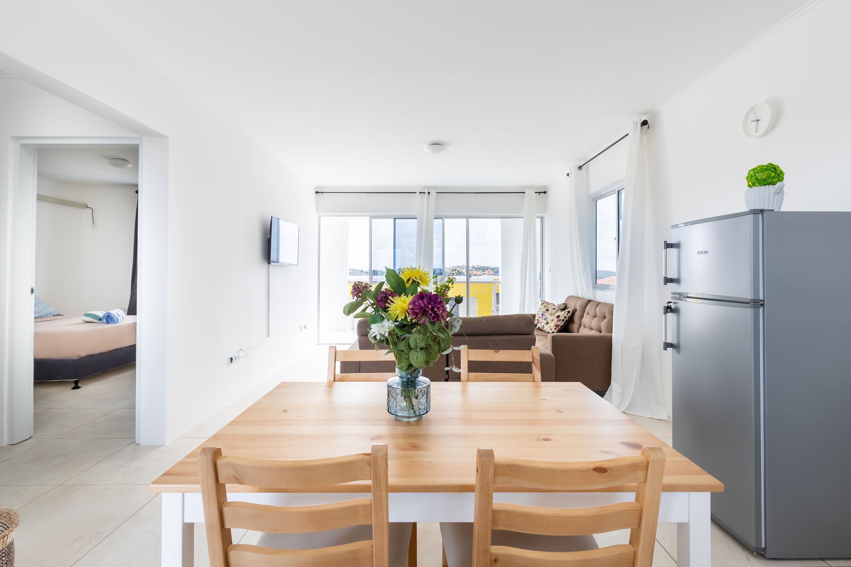 A modern kitchen with a wooden dining table, chairs, a refrigerator, and a window with curtains.