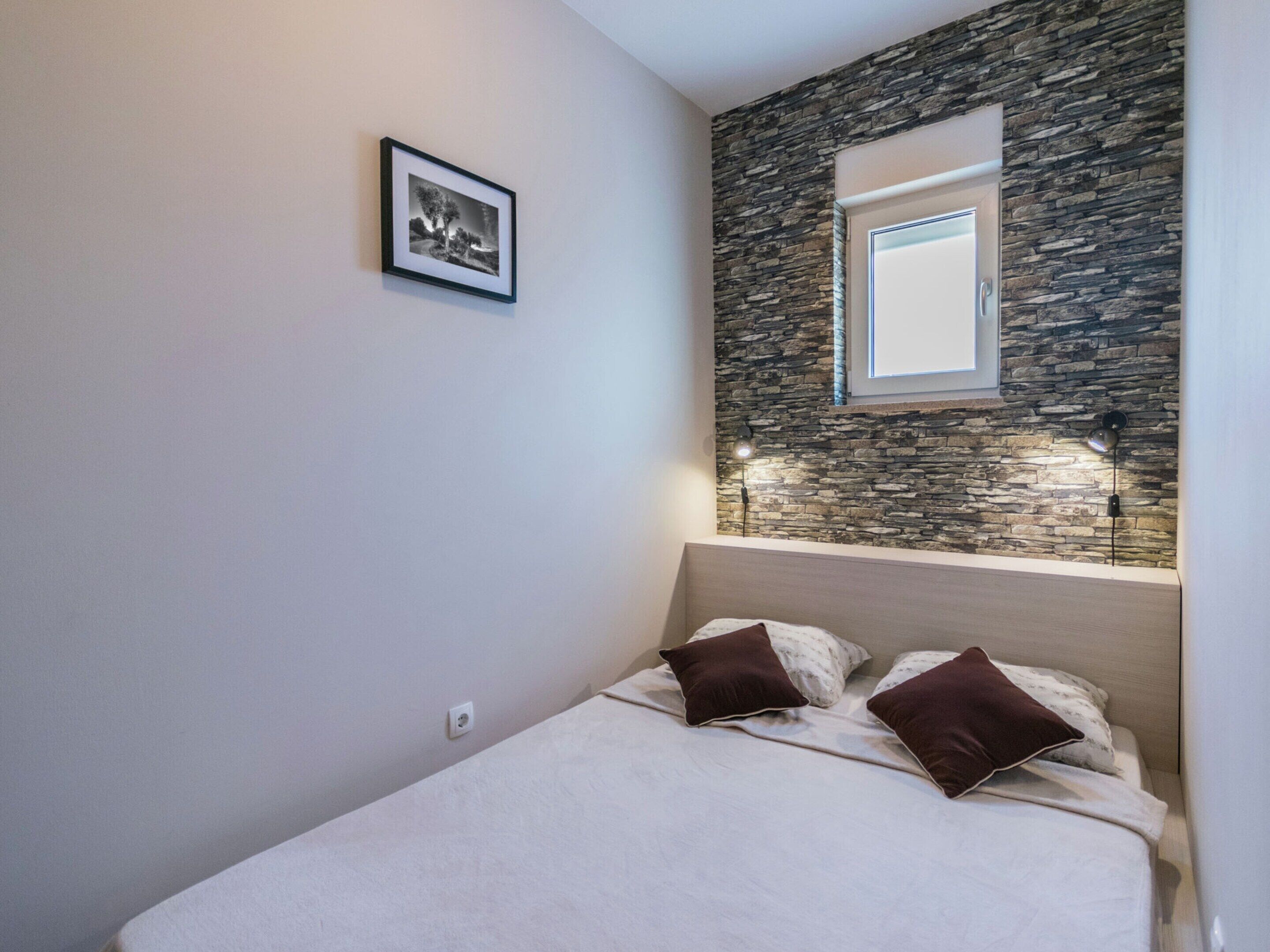 A bedroom with a bed, two pillows, a framed picture on the wall, and a window with a stone wall behind it.