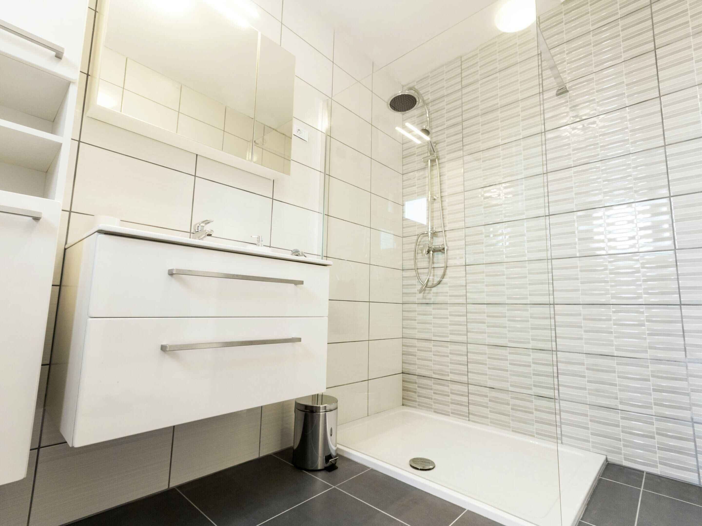 Een moderne badkamer met witte betegelde wanden, een glazen douchecabine en een wit wastafelmeubel met lades.