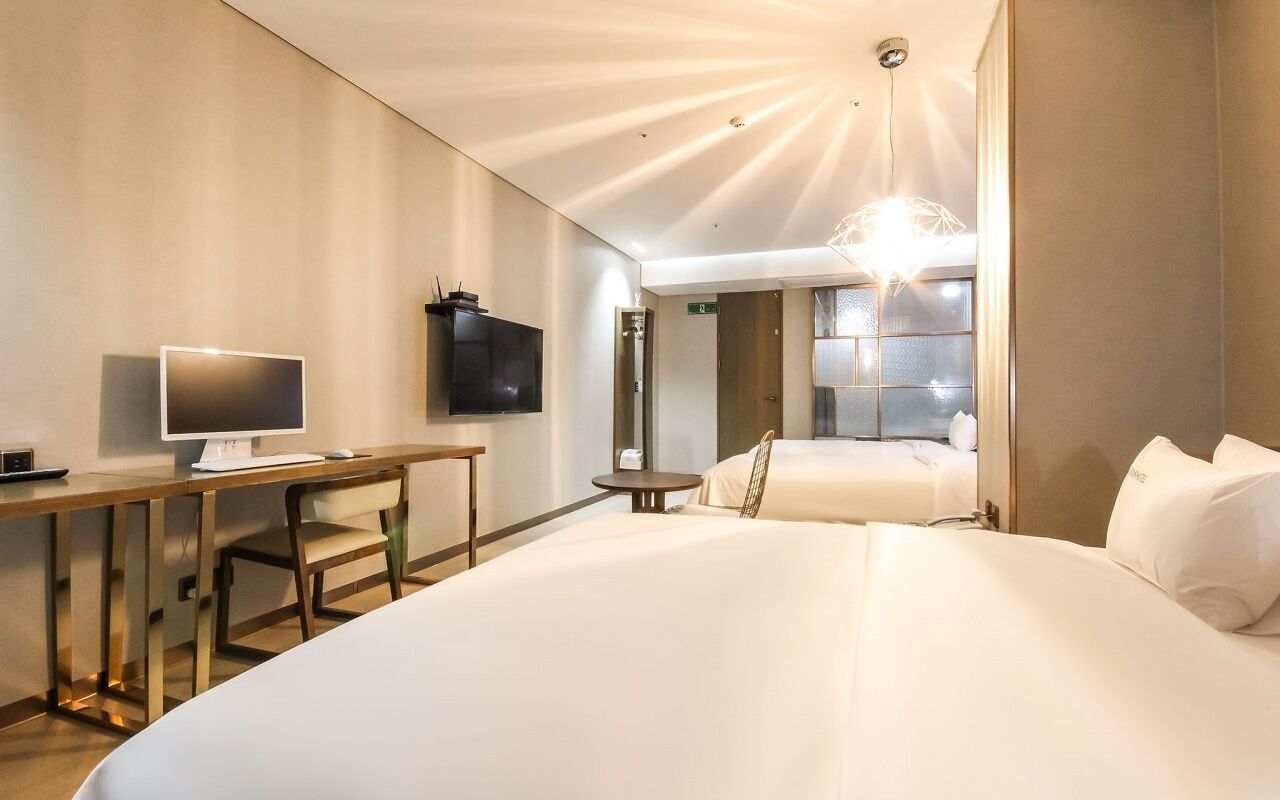 A hotel room with a large bed, a desk with a computer, a TV, and a modern chandelier.