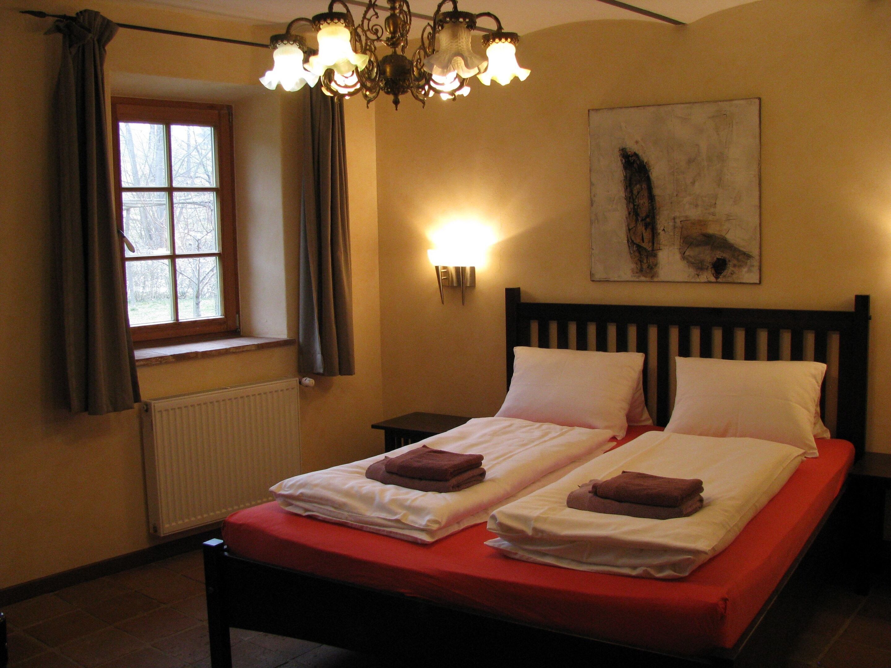 A hotel room with a double bed, a window with curtains, a radiator, a wall-mounted light, and a painting.