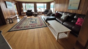 Un salon chaleureux doté d’un grand plancher en bois, d’un canapé en cuir noir, d’un tapis à motifs et offrant une vue sur l’extérieur grâce à de grandes fenêtres.
