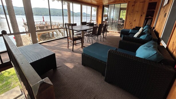 Une cabane en bois dotée d’une grande fenêtre donnant sur un lac. À l’intérieur, on trouve une table à manger avec des chaises, un canapé avec des coussins et un banc en bois.