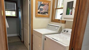 Une buanderie équipée d’un lave-linge et d’un sèche-linge à chargement frontal, d’une porte en bois, d’une fenêtre avec des stores et d’une œuvre d’art encadrée sur le mur, dont le thème est la plage.