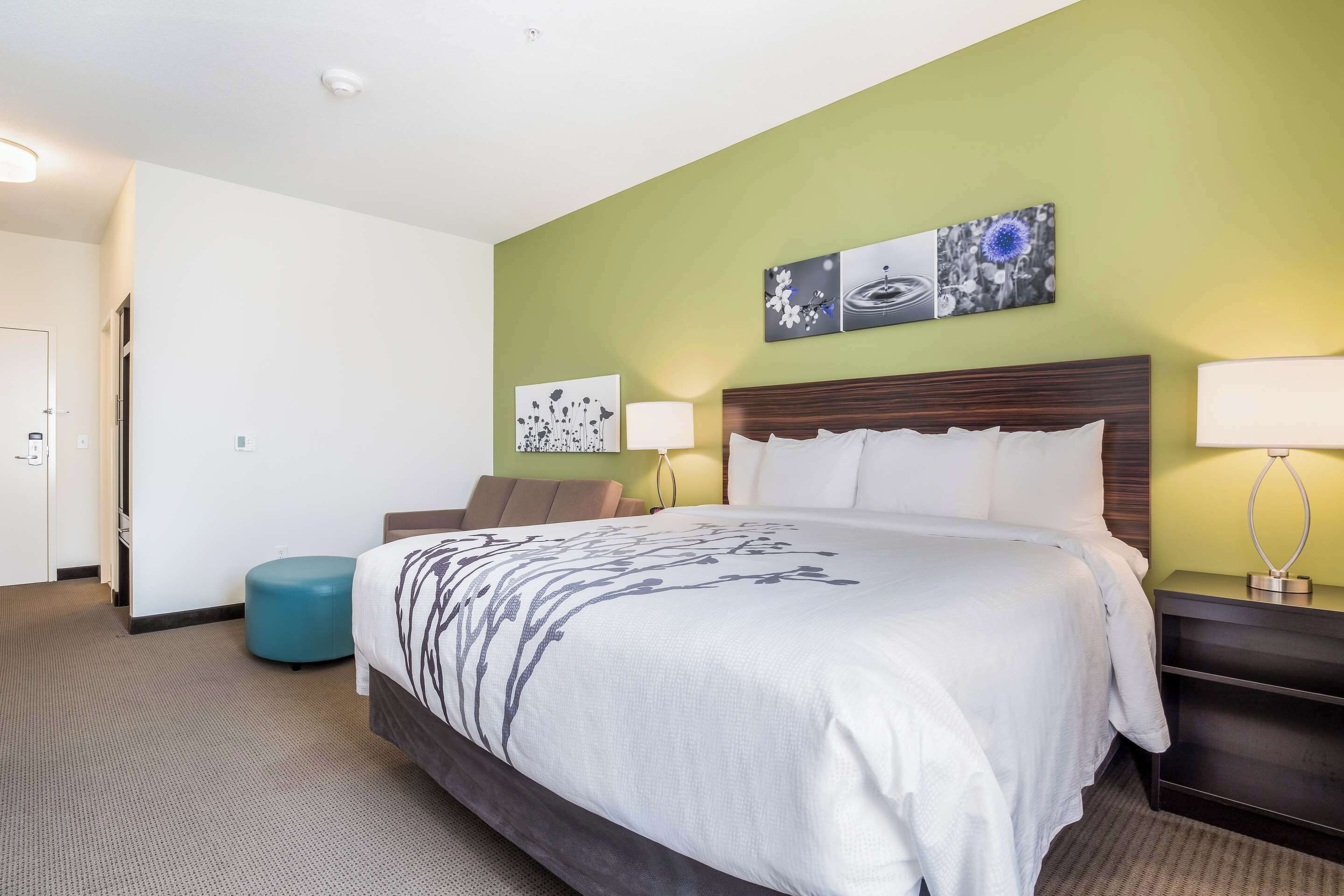 Une chambre d’hôtel avec un grand lit, une table de chevet avec une lampe, une œuvre d’art accrochée au mur et un pouf de couleur turquoise.