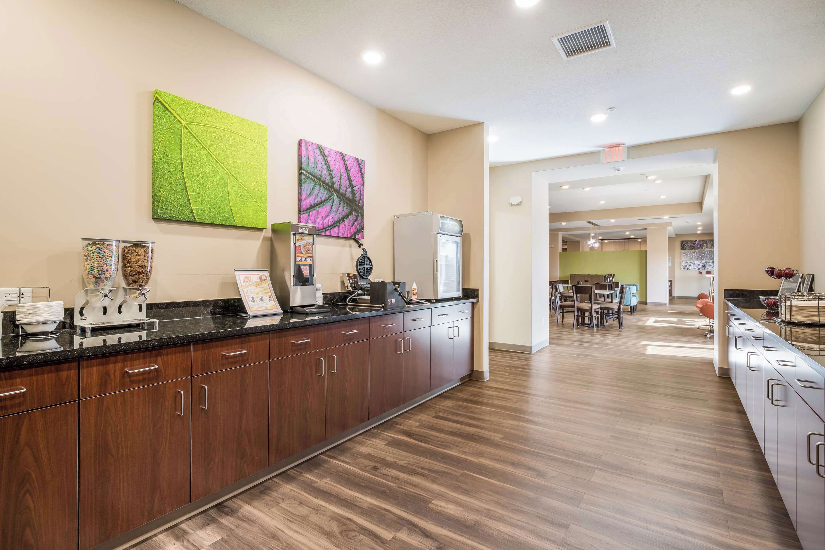 A modern kitchen with wooden cabinets, a black countertop, and a refrigerator. There are two green leaf paintings on the wall and a dining area in the background.